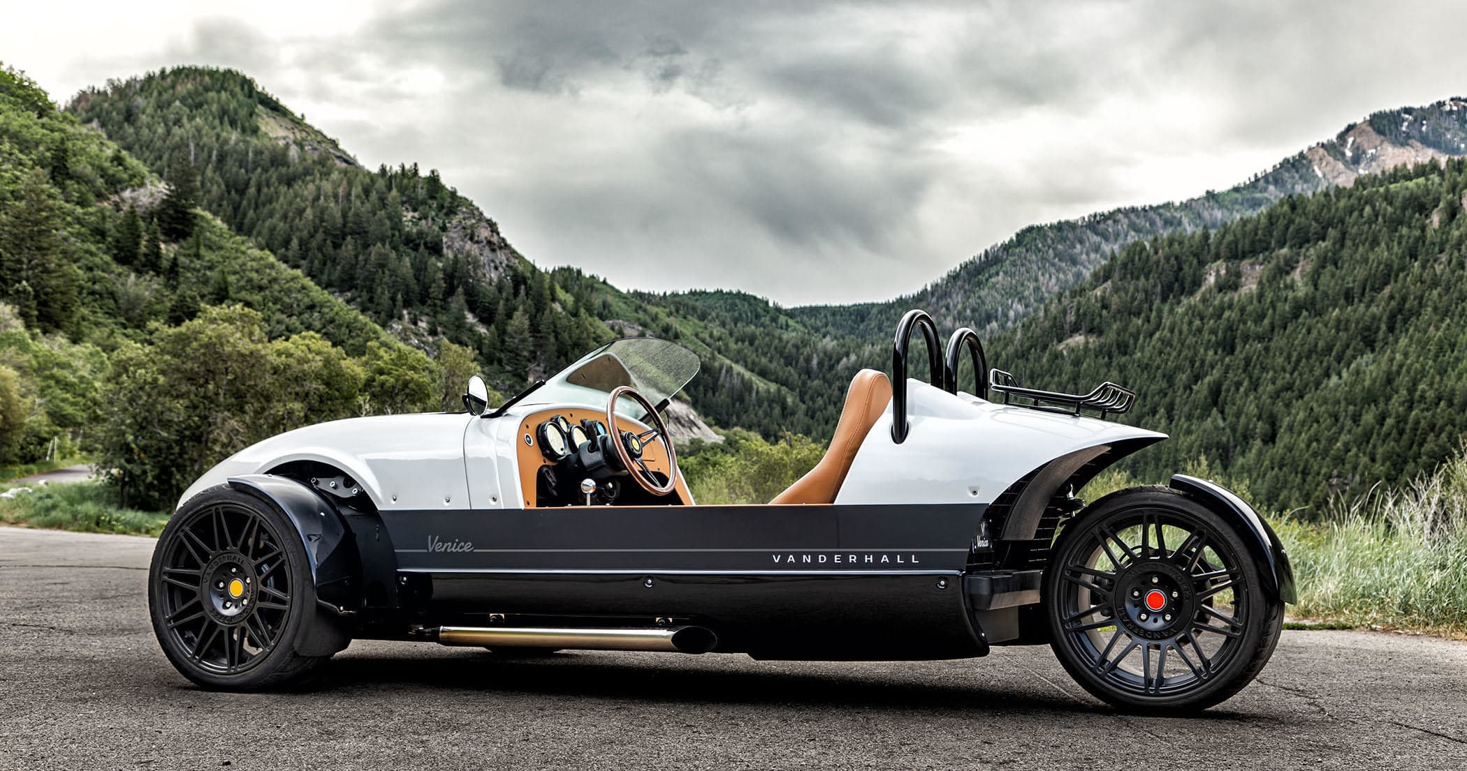 Open-top three-wheeled sports car on scenic alpine road near mountains