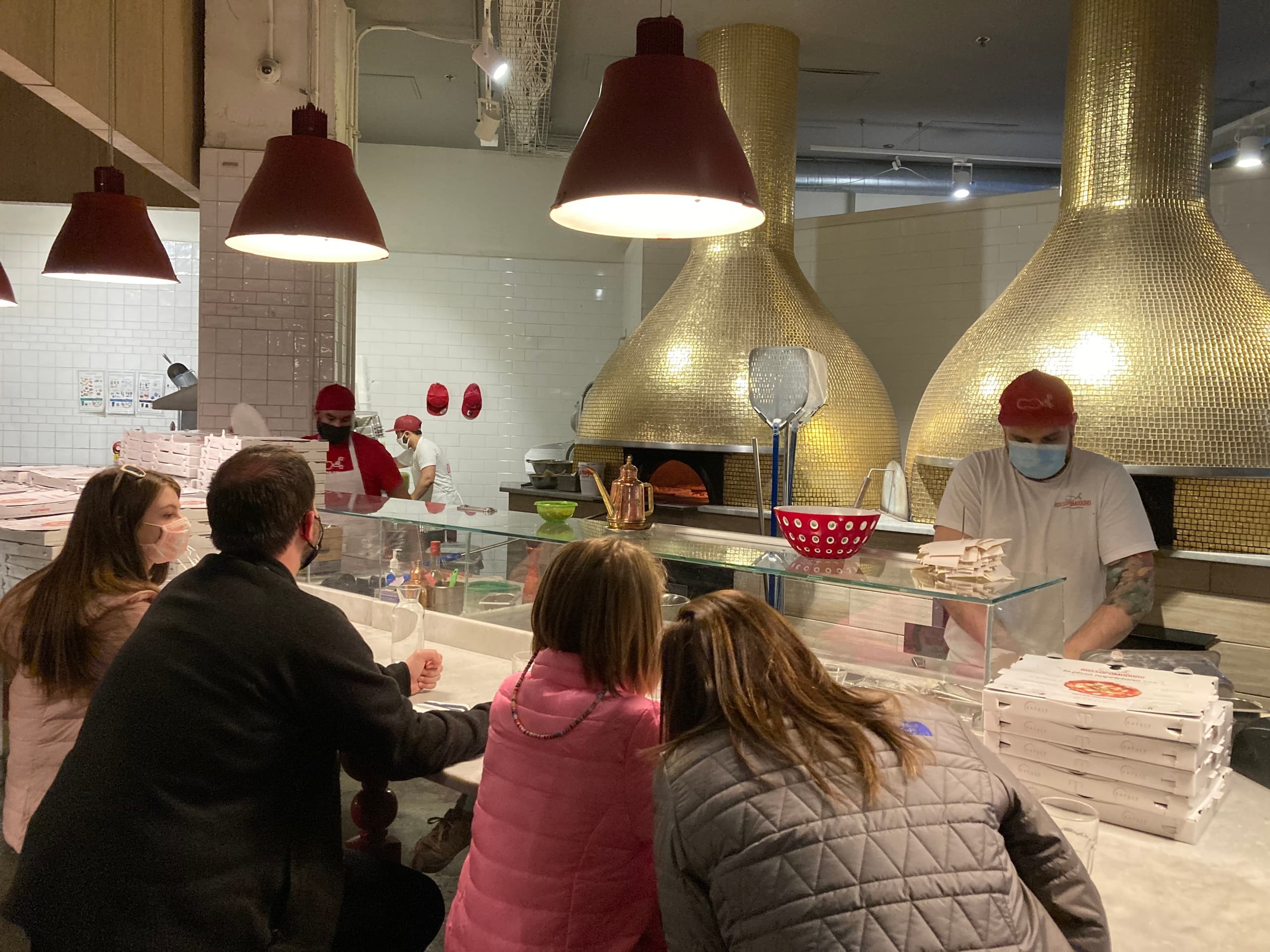 Customers ordering pizza at indoor pizzeria counter with ovens