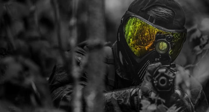 Masked paintball combatant peeking through foliage in woods