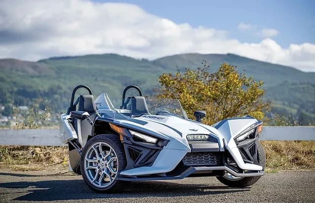 Open-air three-wheeled roadster parked by lakeside mountain road
