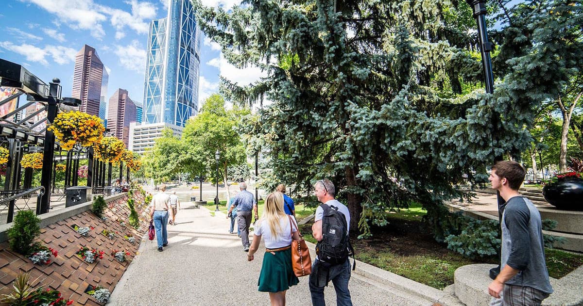 People strolling along a sunny urban park path with Calgary skyline