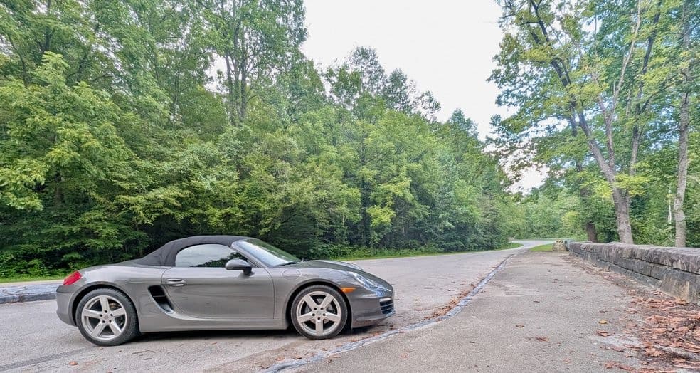 Silver Porsche convertible side on tree-lined road scenic cruise