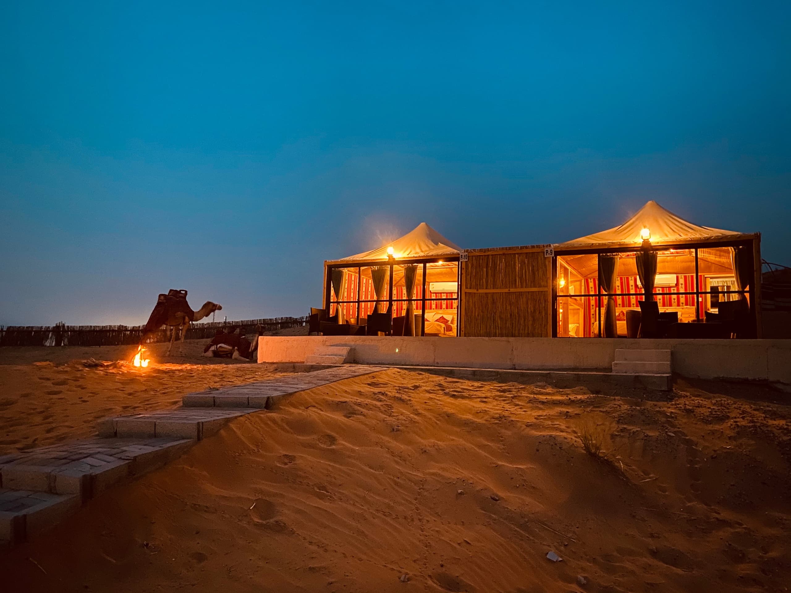 Lit desert camp tents at dusk with camel and campfire