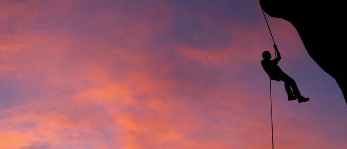 Silhouette climber rappelling off cliff at sunset over sky