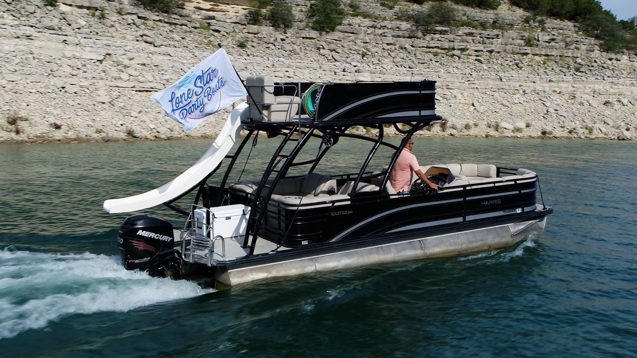 Black pontoon boat powering past rocky shore on clear lake