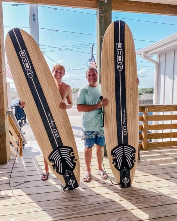 Two surfers holding longboards on beach house deck, ready