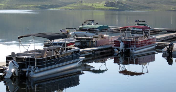 Multiple pontoons docked on reflective lake, green hills backdrop