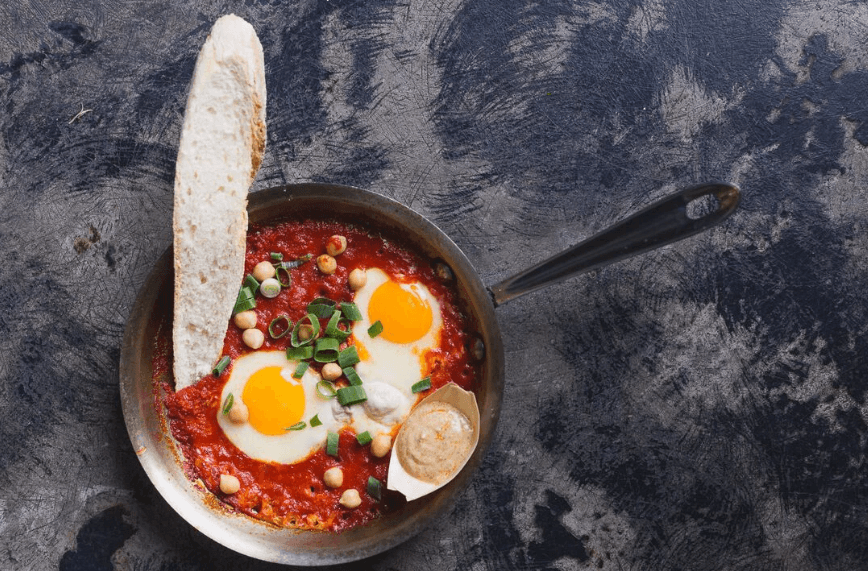 Skillet shakshuka with eggs, bread and herbs in rustic kitchen