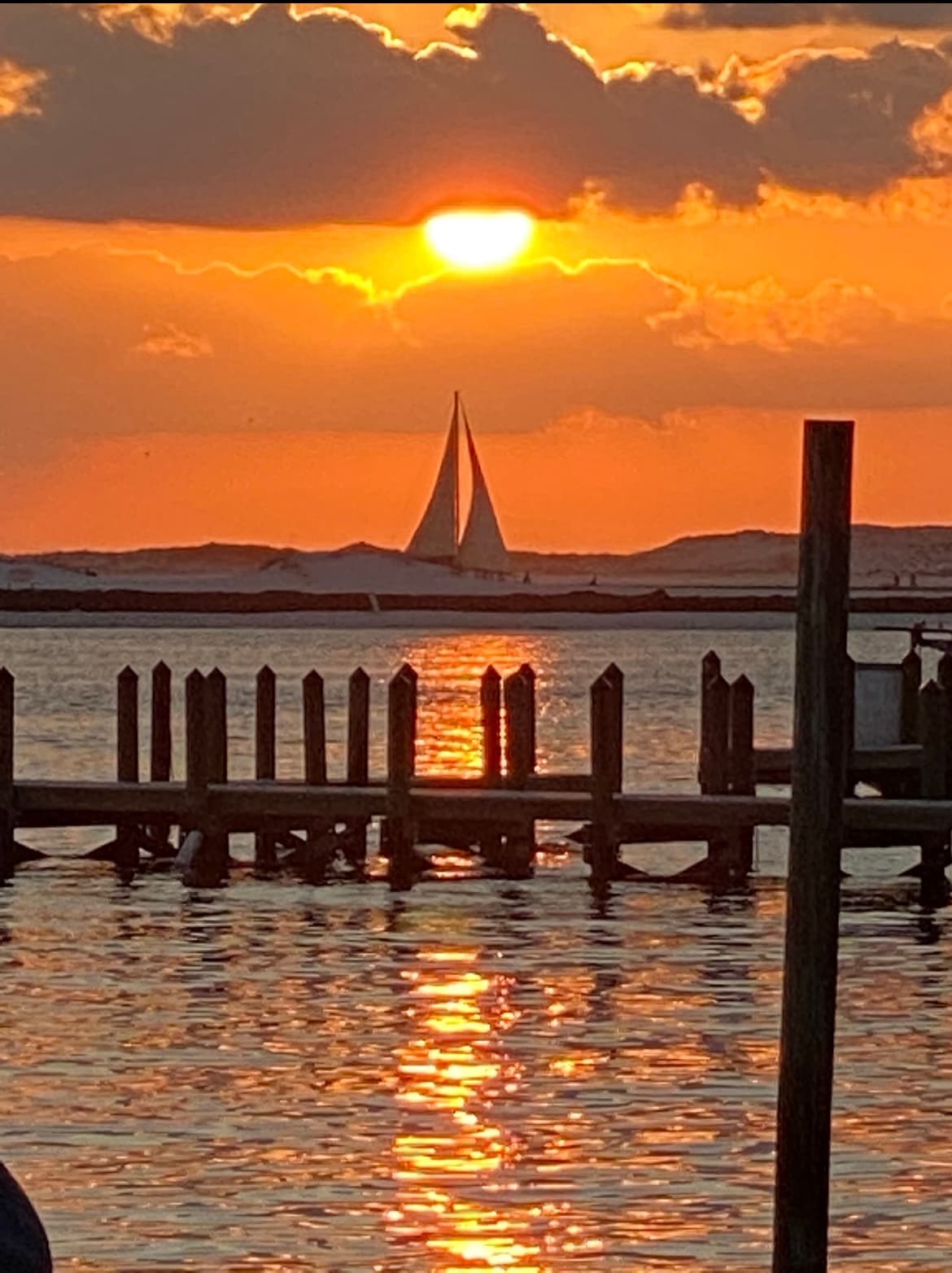 Sailboat silhouette at sunset behind pier, calm coastal harbor view