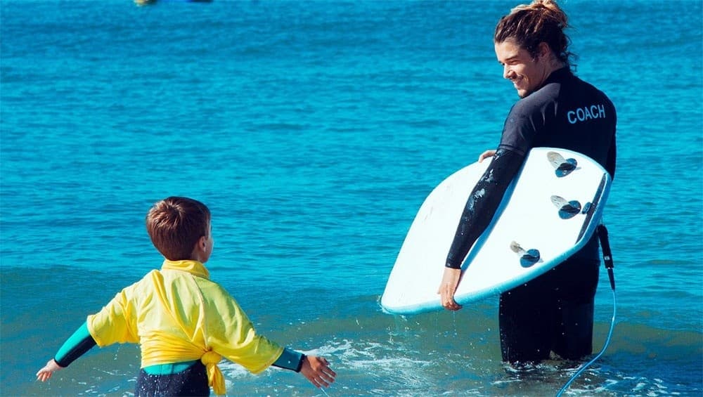 Surf coach assisting child with board in shallow Carcavelos sea