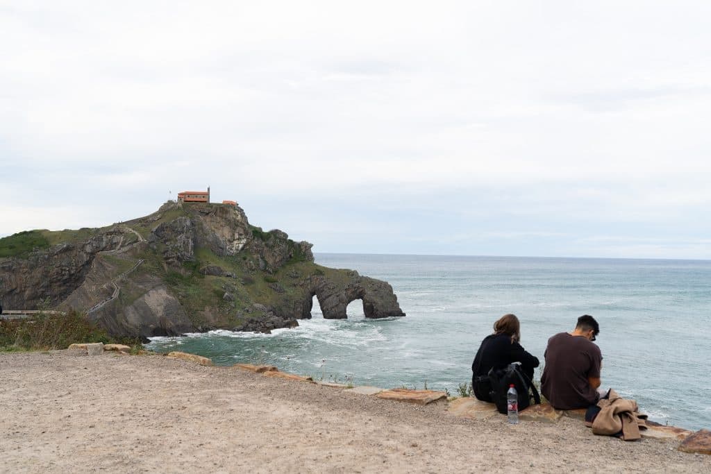 Couple seated on coastal cliff overlooking sea arch, coastal hiking view
