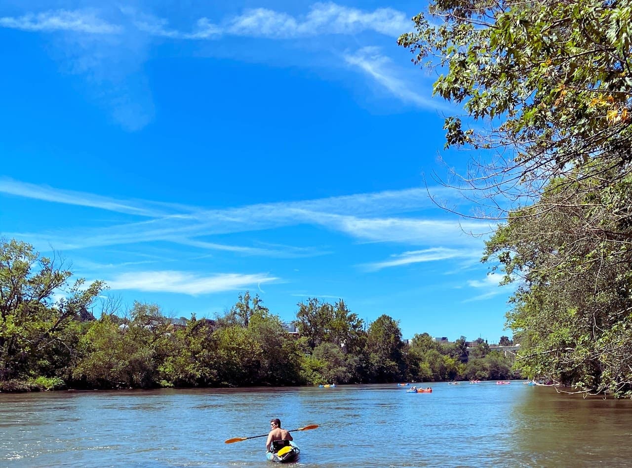 Solo kayaker paddling down scenic tree-lined river under blue sky