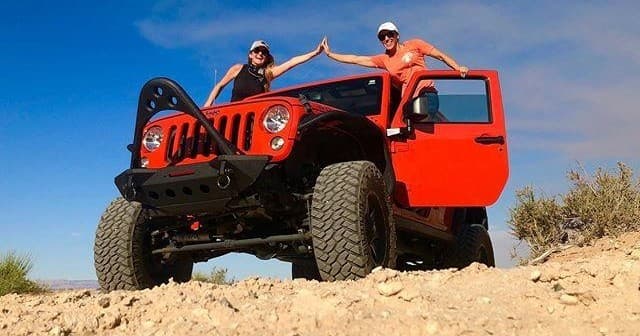 Friends high-fiving on lifted Jeep over rocky desert hill under clear sky