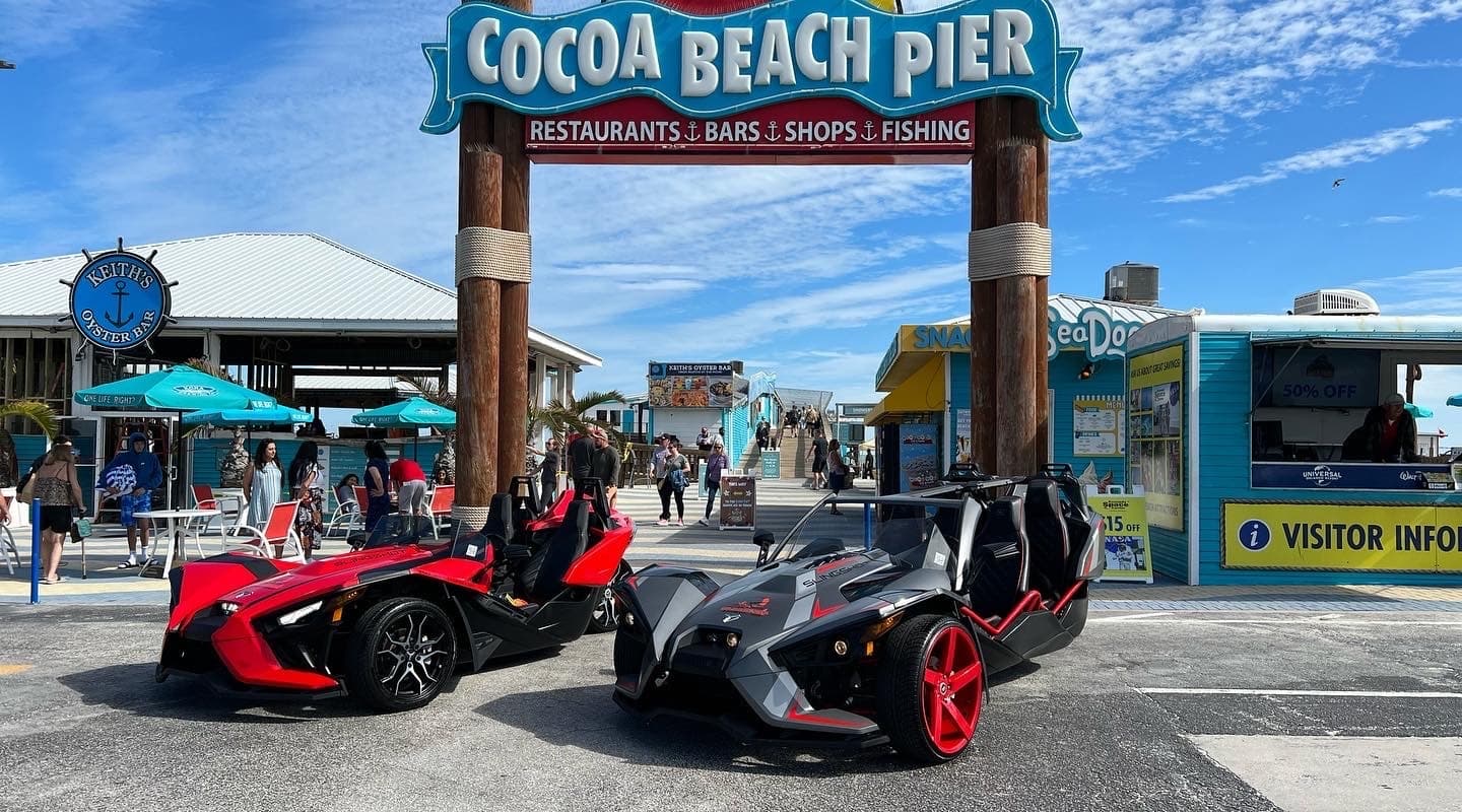 Two slingshot roadsters parked at Cocoa Beach Pier entrance