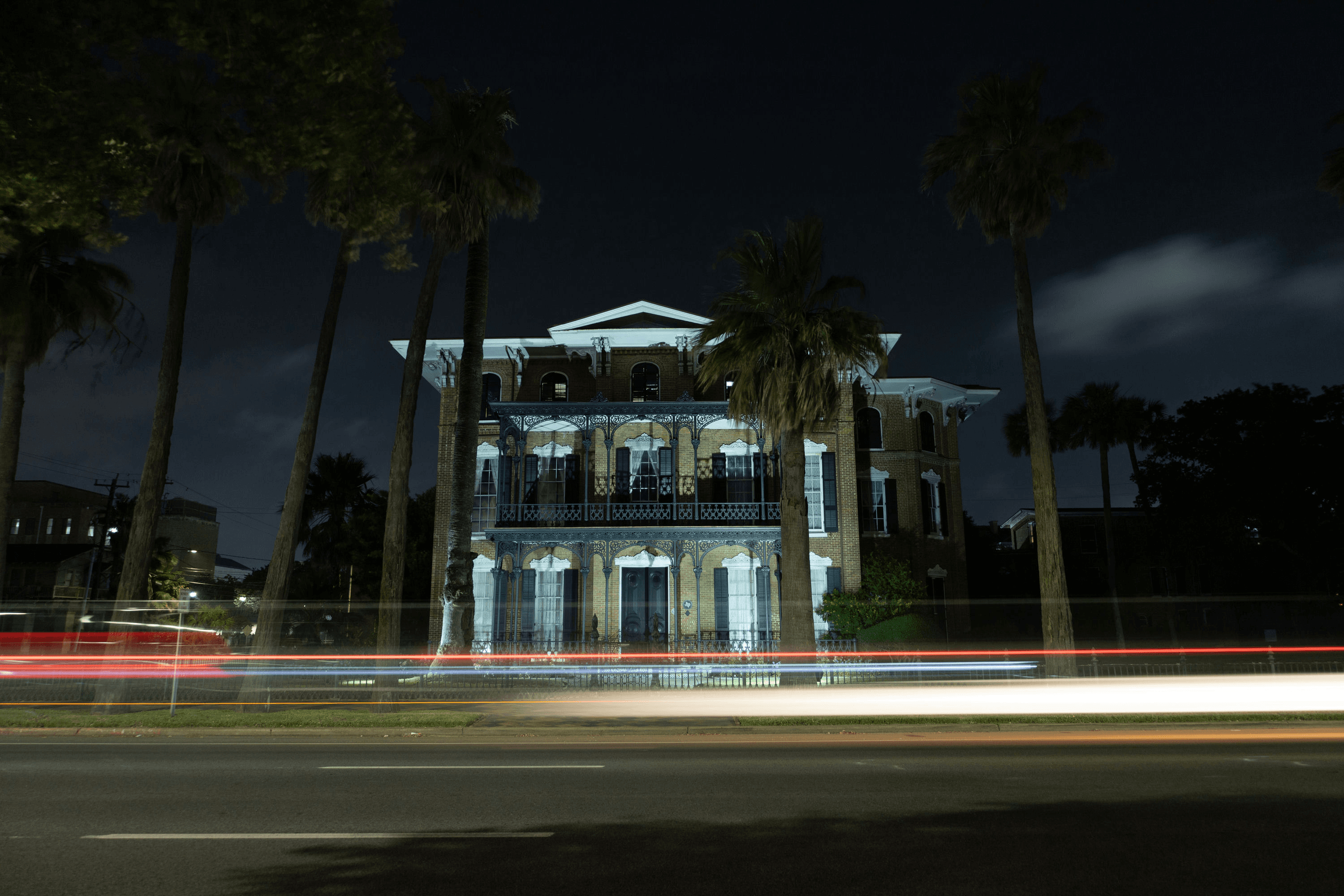 Night long-exposure of mansion on palm-lined street with car light trails