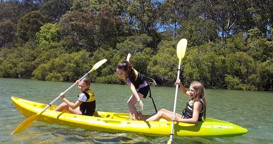 Three kids paddling a tandem yellow kayak on a quiet estuary