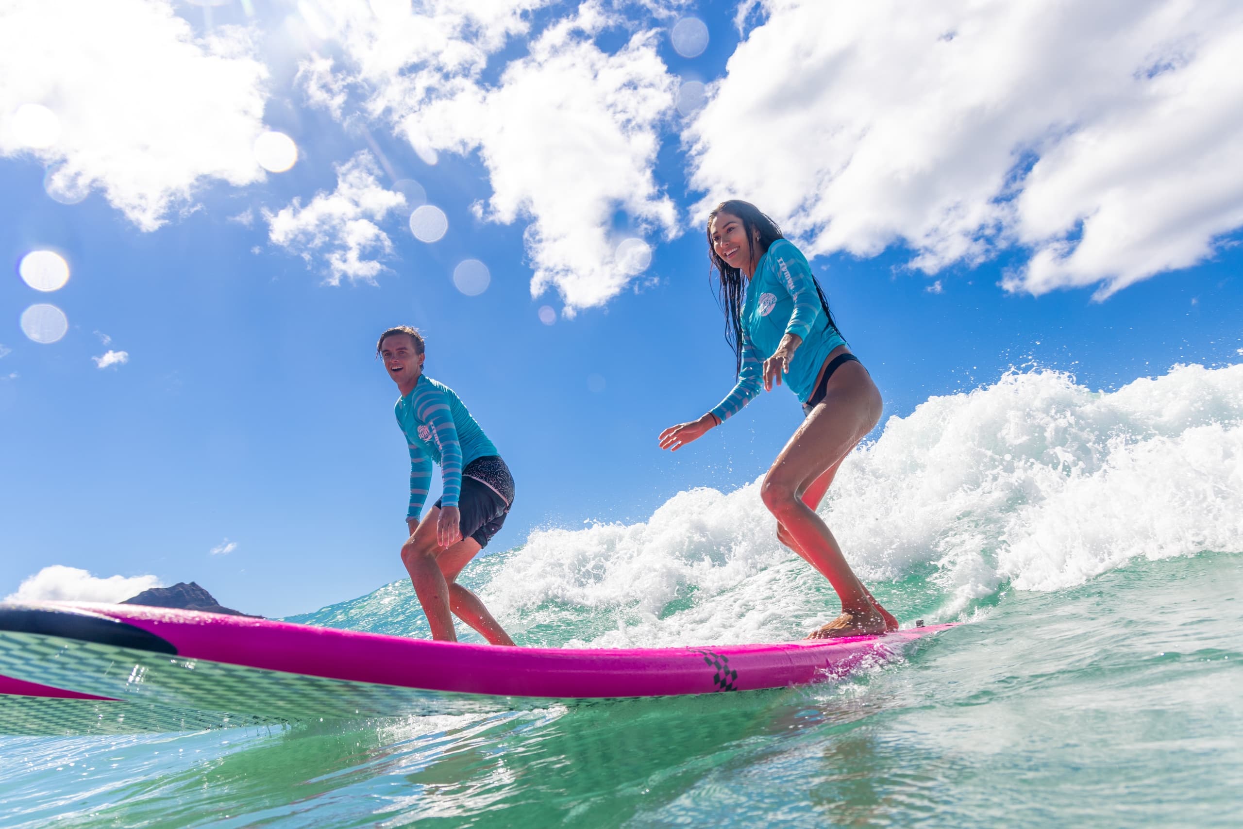 Two surfers balancing on pink longboard under bright blue sky