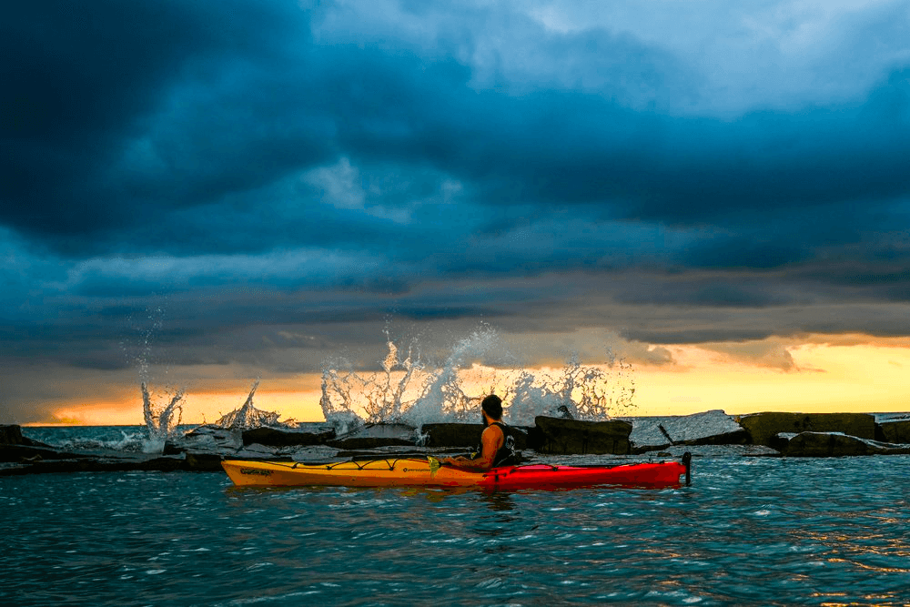 Solo kayaker splashing near rocky breakwater under dramatic sunset skies
