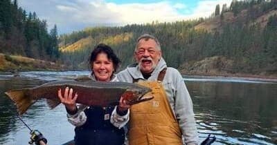 Happy anglers holding large freshwater trout on riverbank, forested hills