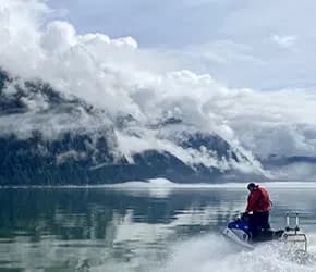 Jet skier cruising reflective fjord with cloud-shrouded Alaskan mountains
