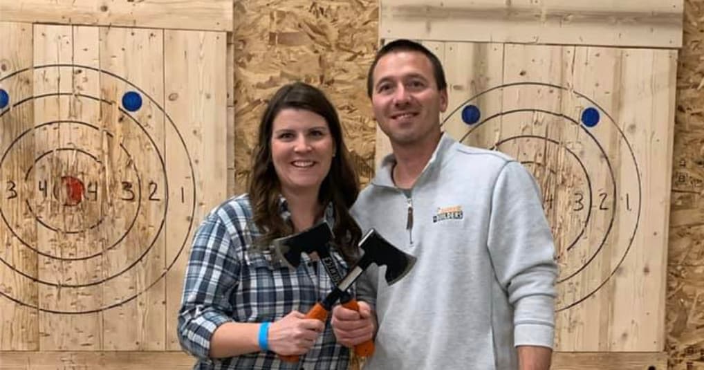 Couple holding axes in front of indoor axe throwing wooden targets