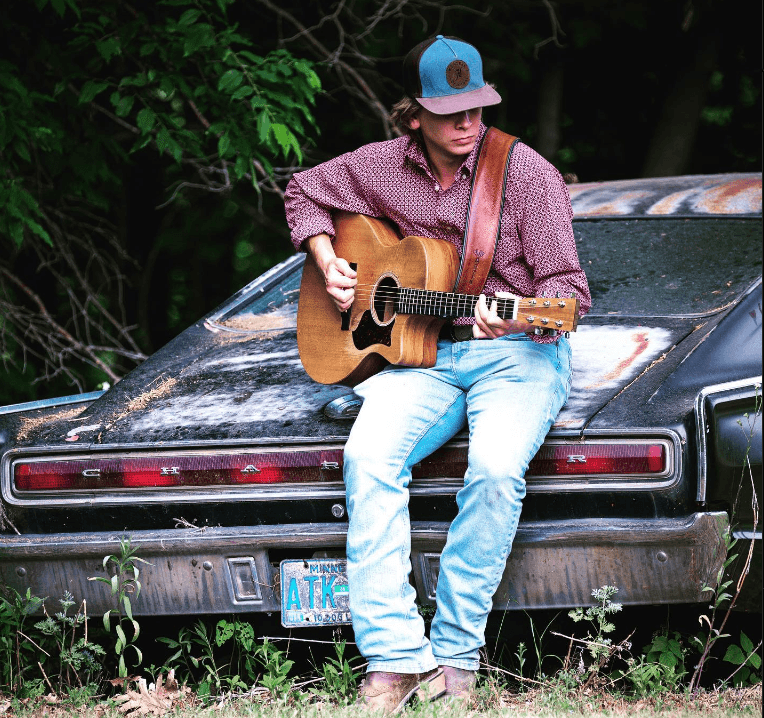 Young man playing acoustic guitar sitting on old car outdoors
