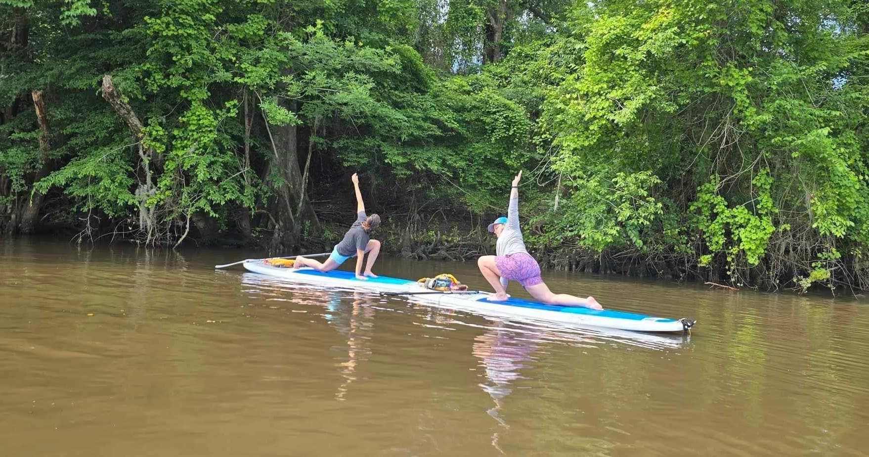 Two people doing paddleboard yoga near riverbank with green trees