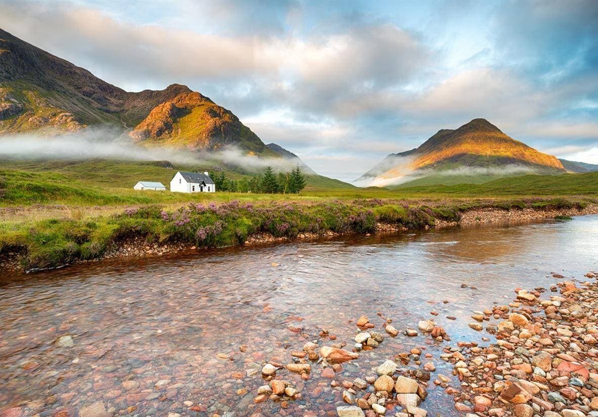 Riverside cottage with misty mountains and pebble stream at dawn