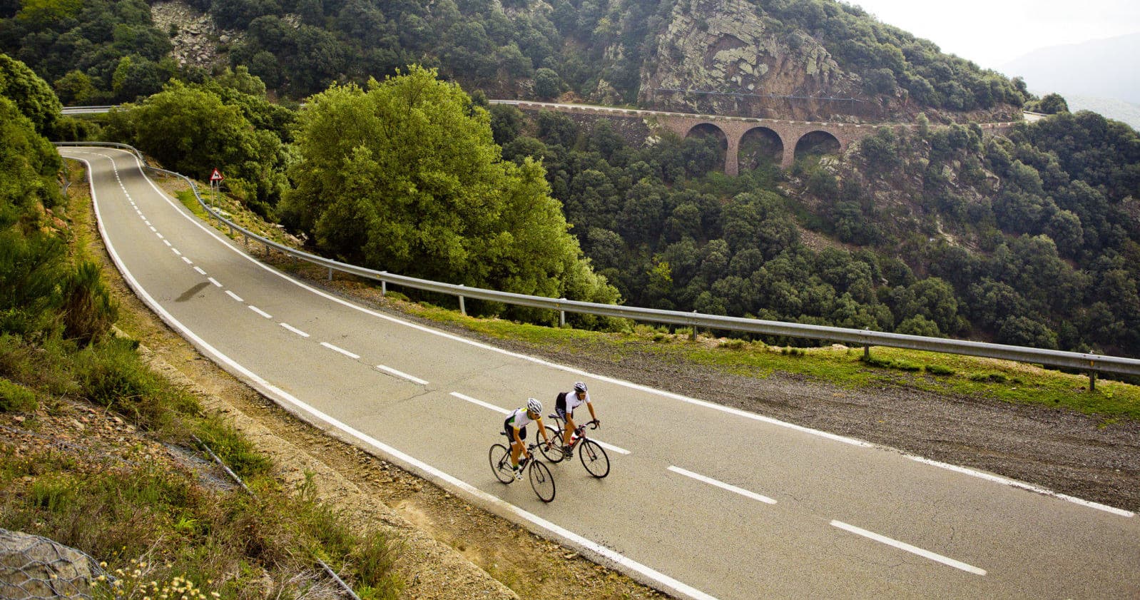 Cyclists on quiet country road under viaduct through green hills