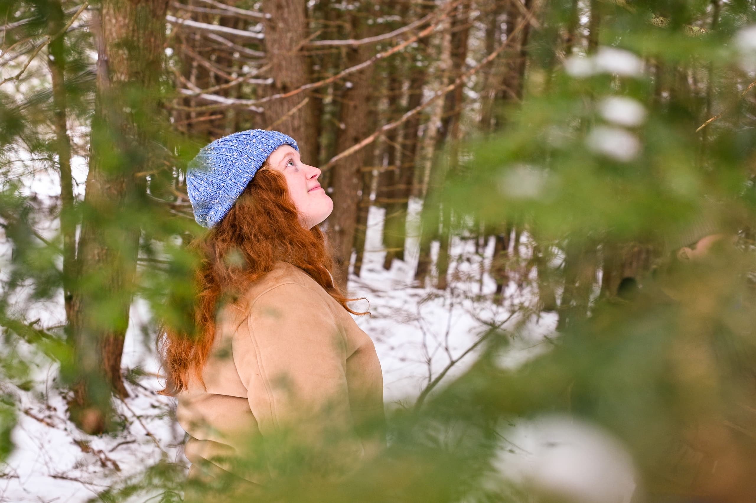 Woman in blue hat hiking in snowy evergreen forest, looking up
