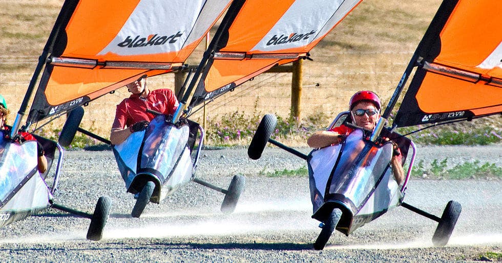 Three land yachts kicking up dust on open gravel beach course