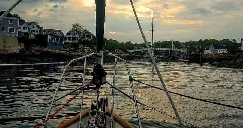Sailboat bow heading toward rocky seaside village at golden sunset