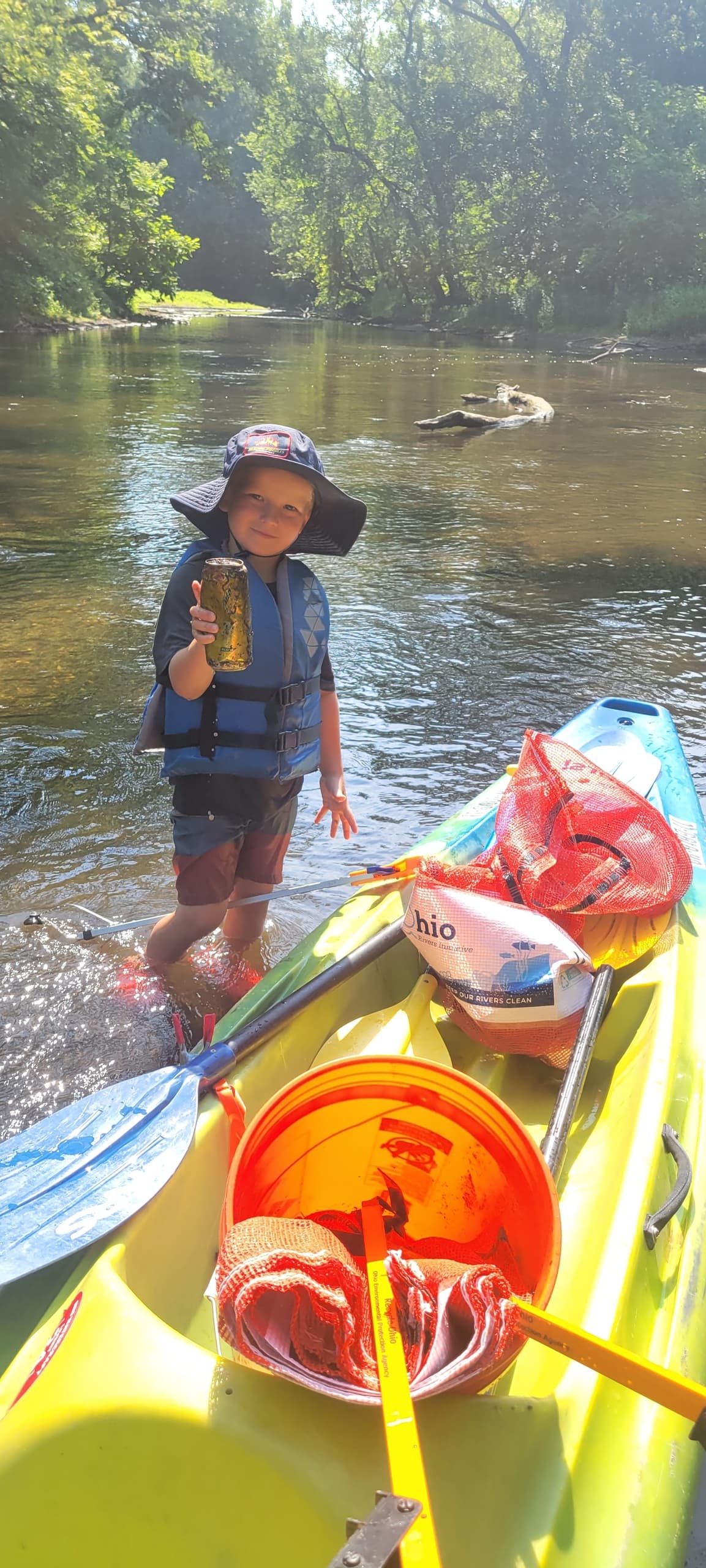 Child beside kayak holding bottle on shallow riverbank adventure