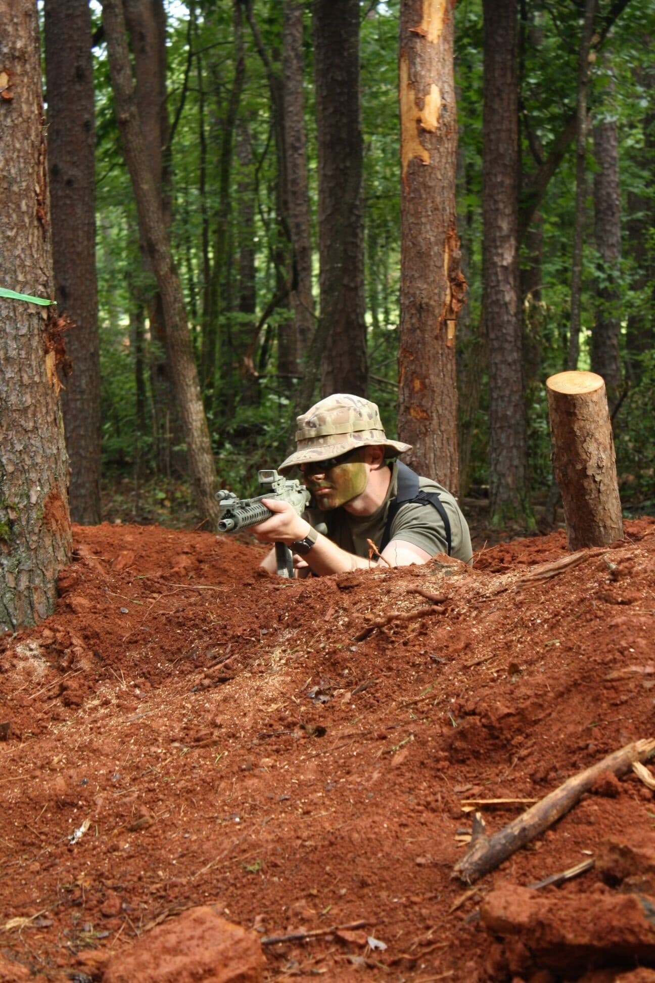 Prone airsoft player aiming from dirt berm in wooded terrain
