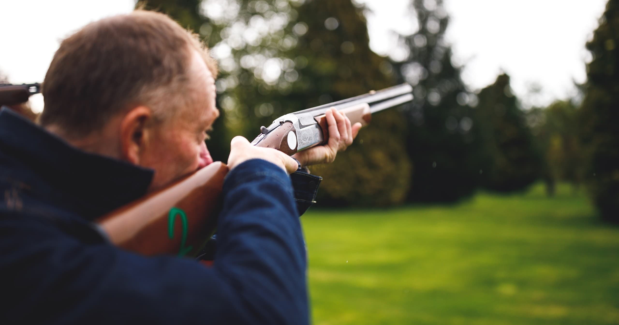 Clay shooter aiming shotgun across open grassy countryside range