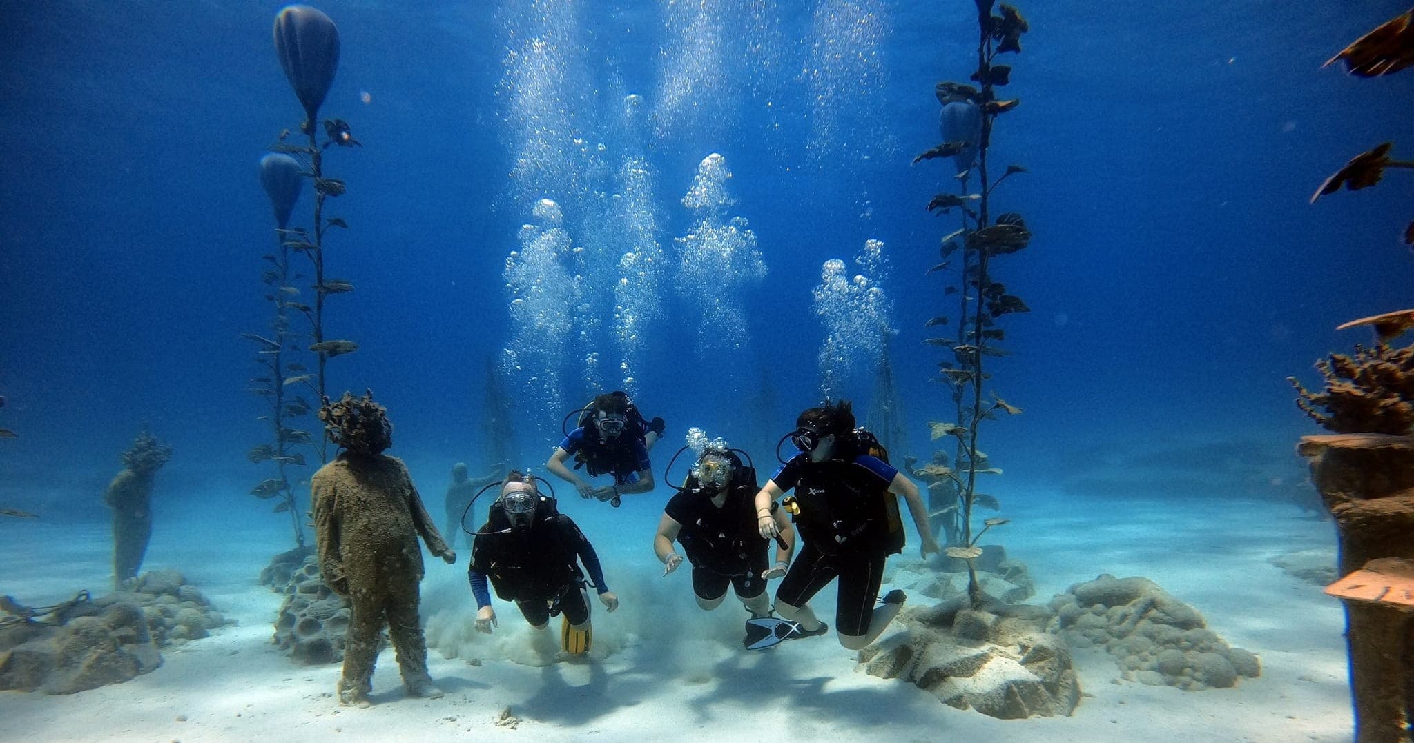Group of scuba divers exploring submerged statues and kelp forest reef