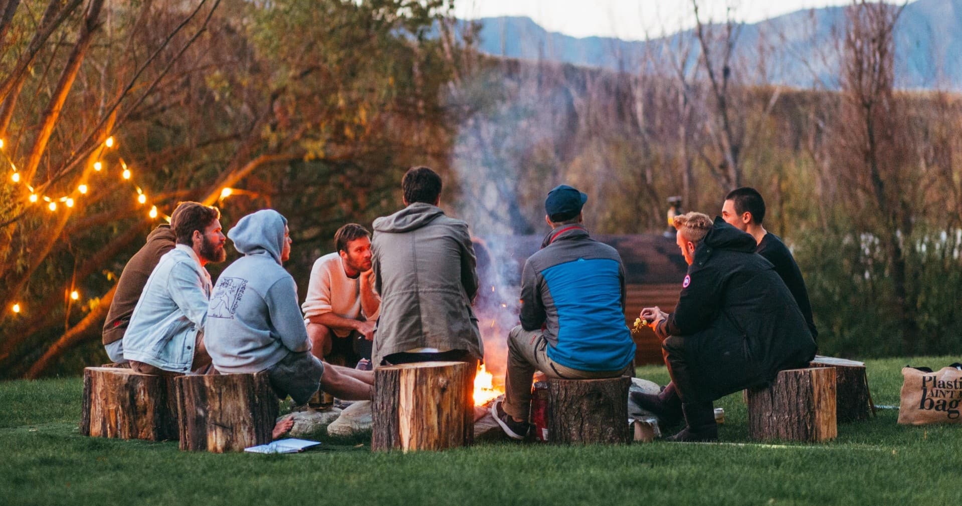 Friends gathered around campfire at dusk in mountain meadow