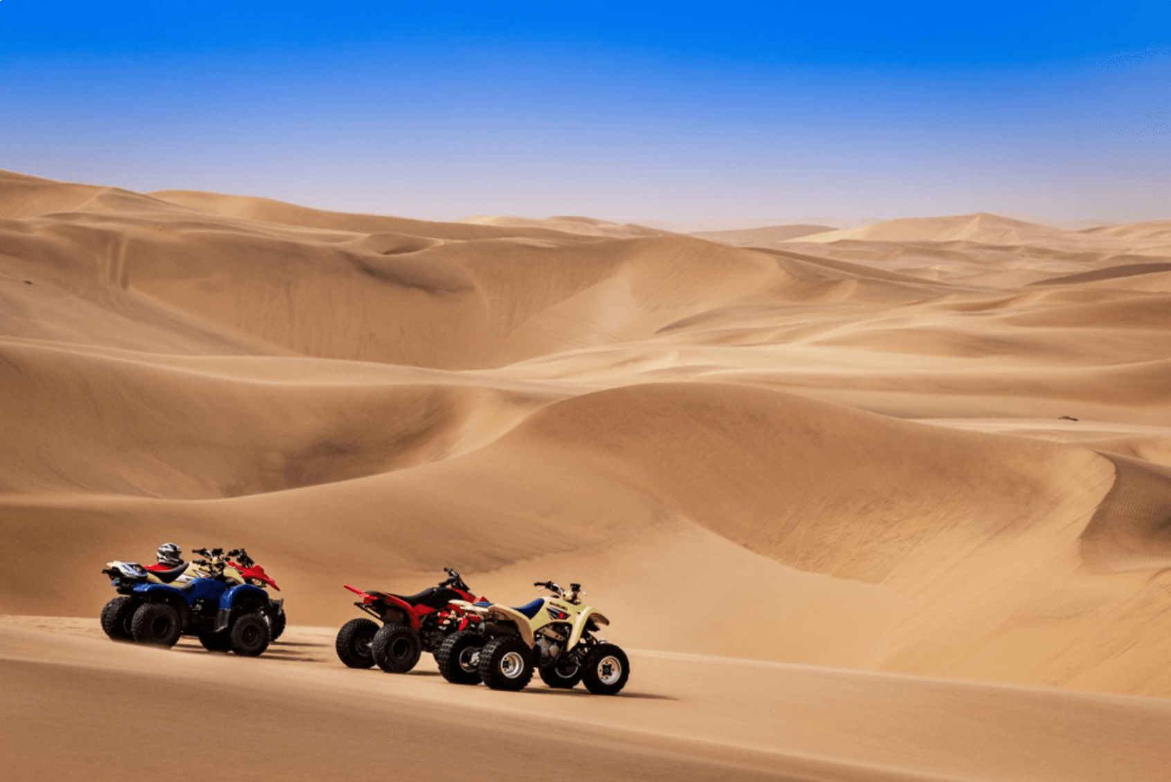 Three ATVs riding across wide desert dunes under blue sky