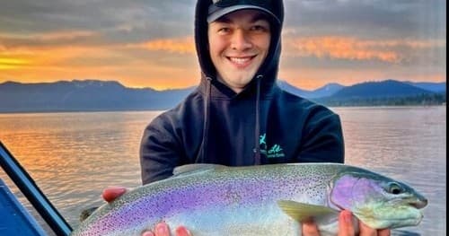 Smiling angler holding rainbow trout on lake at sunset