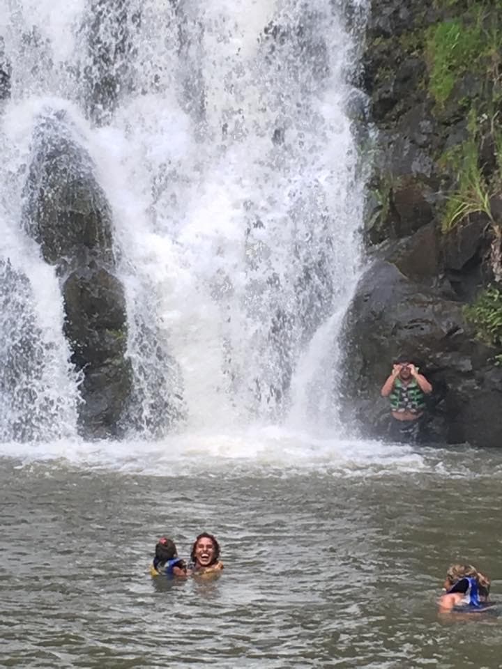 People swimming beneath a cascading waterfall in lush tropical pool