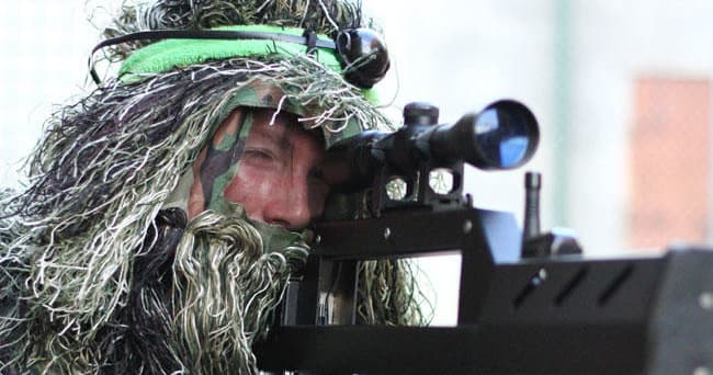 Ghillie-suited player aiming scoped rifle in woodland