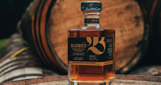 Bladnoch whisky bottle centered against outdoor oak casks on distillery grounds
