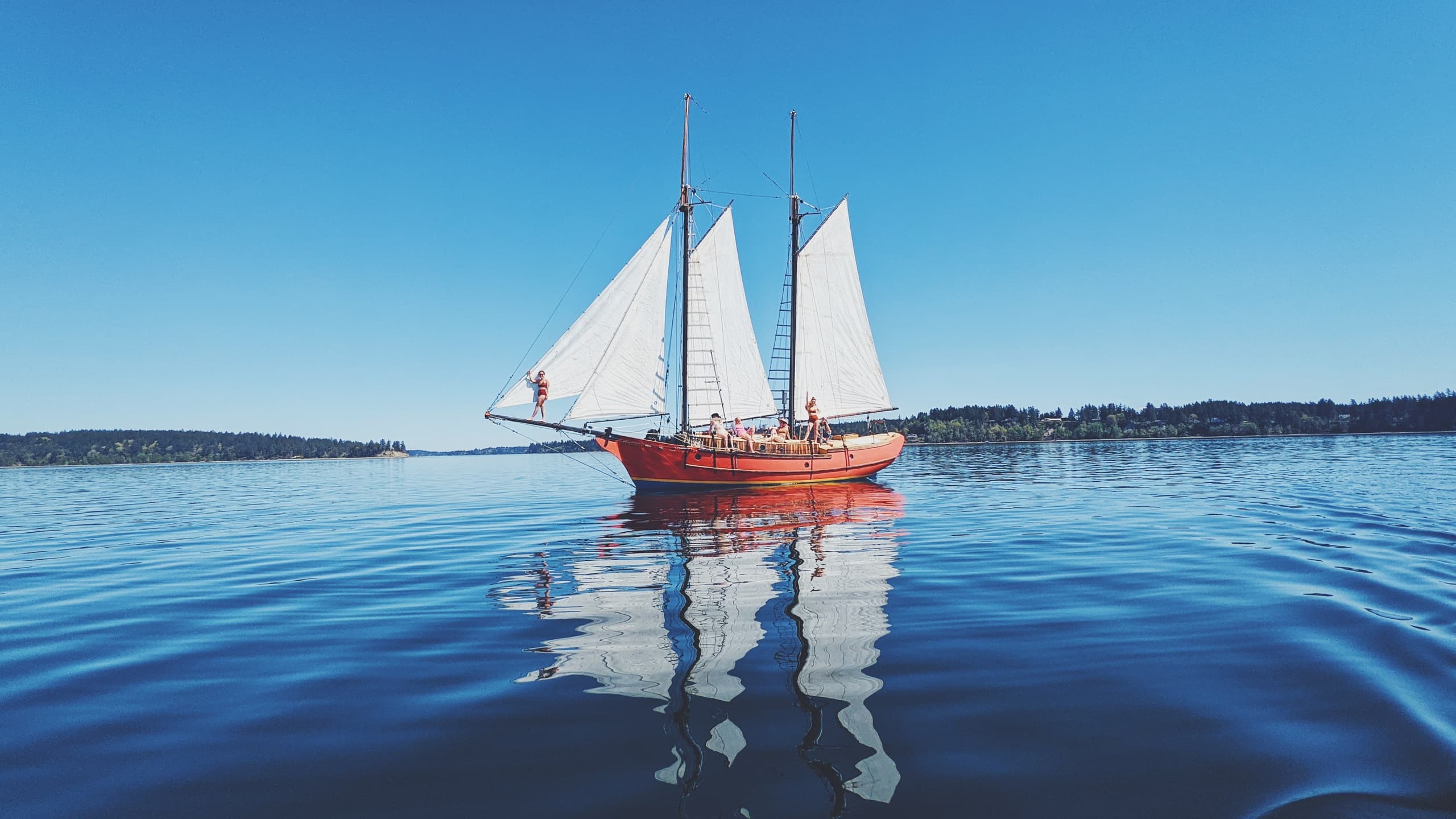 Red schooner sailing on calm coastal waters with reflection