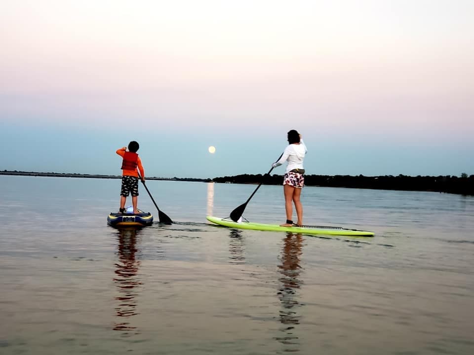 Two paddleboarders at sunrise with moon over bay