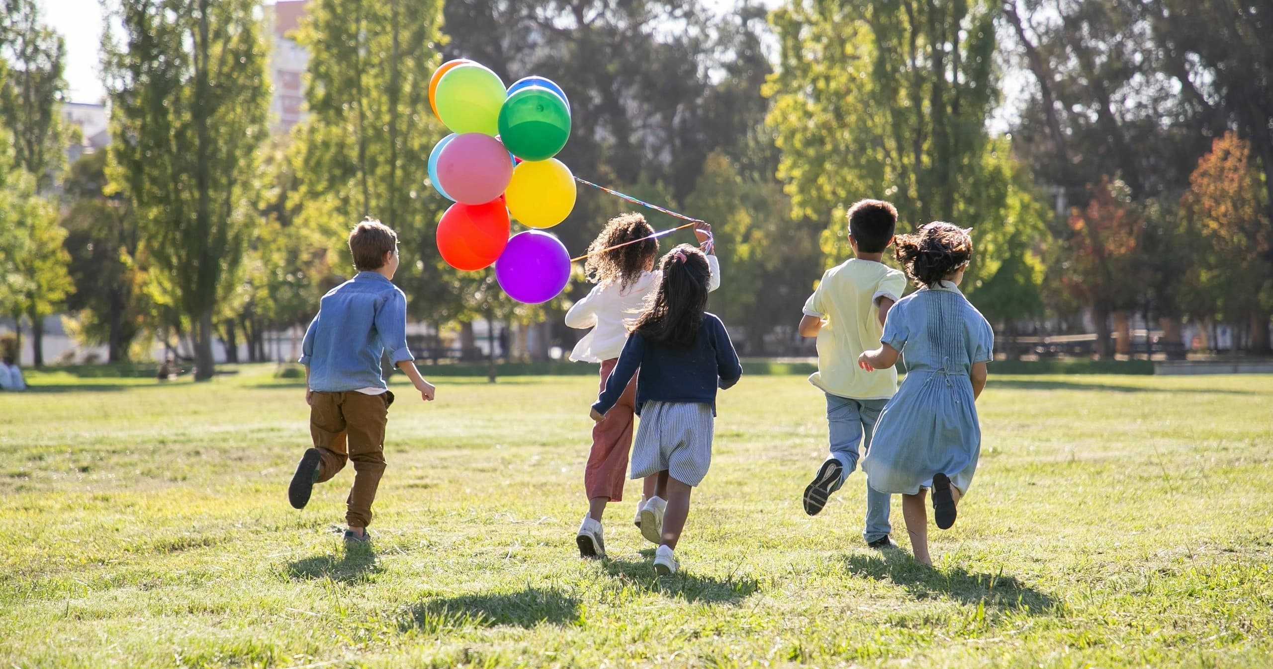 Children running across sunny park meadow holding colorful balloons
