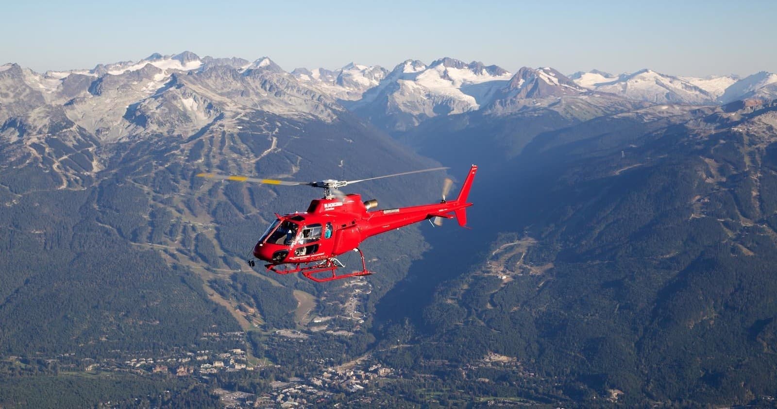 Red helicopter mid-flight over jagged snow-capped mountain peaks