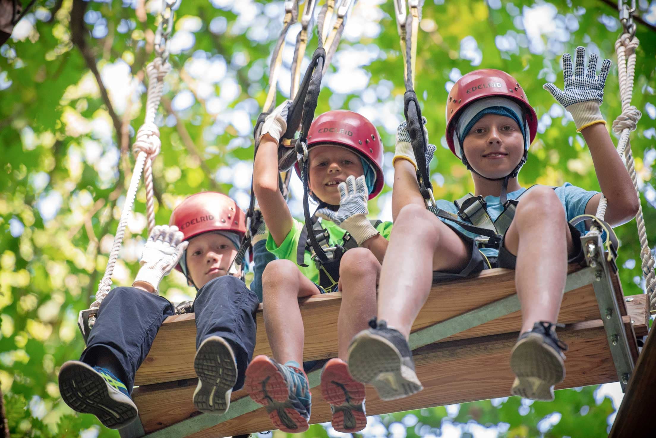 Children in helmets on suspended wooden platform waving in forest