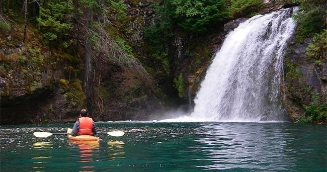 Paddler in kayak approaching waterfall in lush green gorge