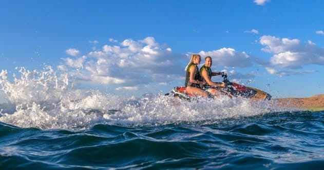 Two people jet skiing across choppy coastal surf