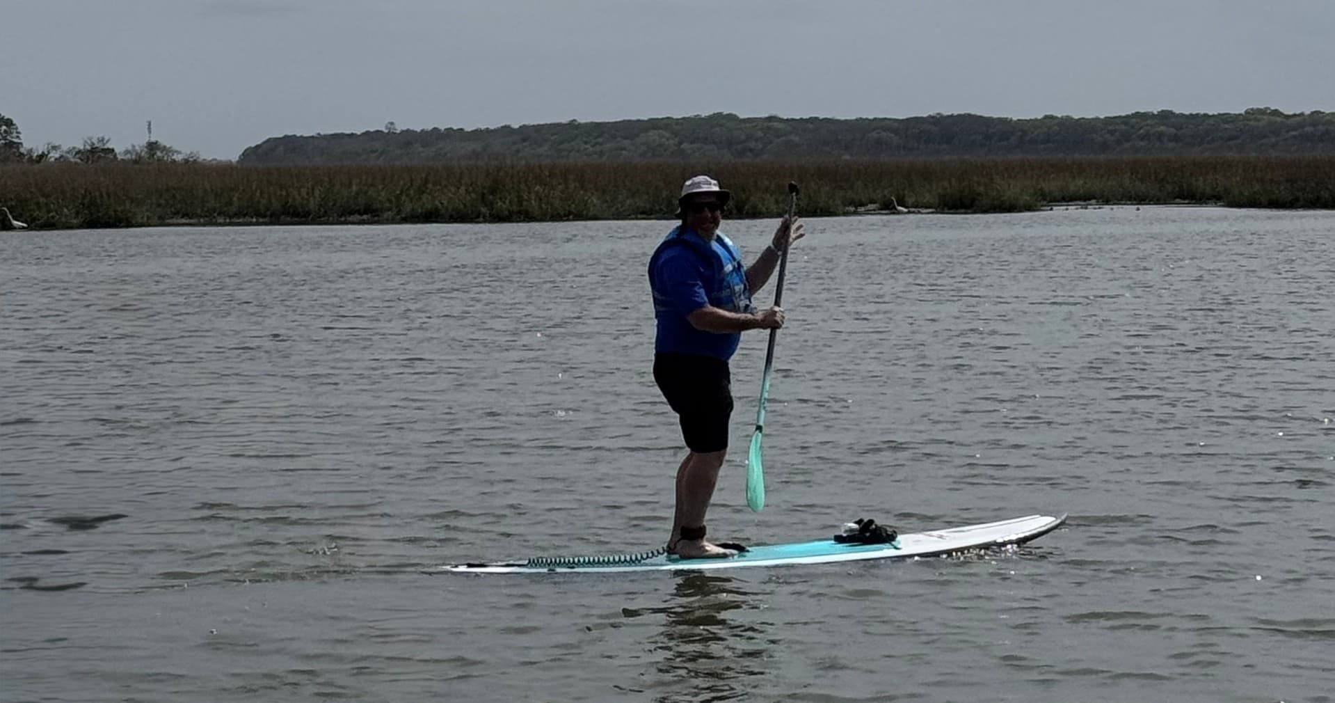 Stand-up paddleboarder paddling across open gray tidal creek